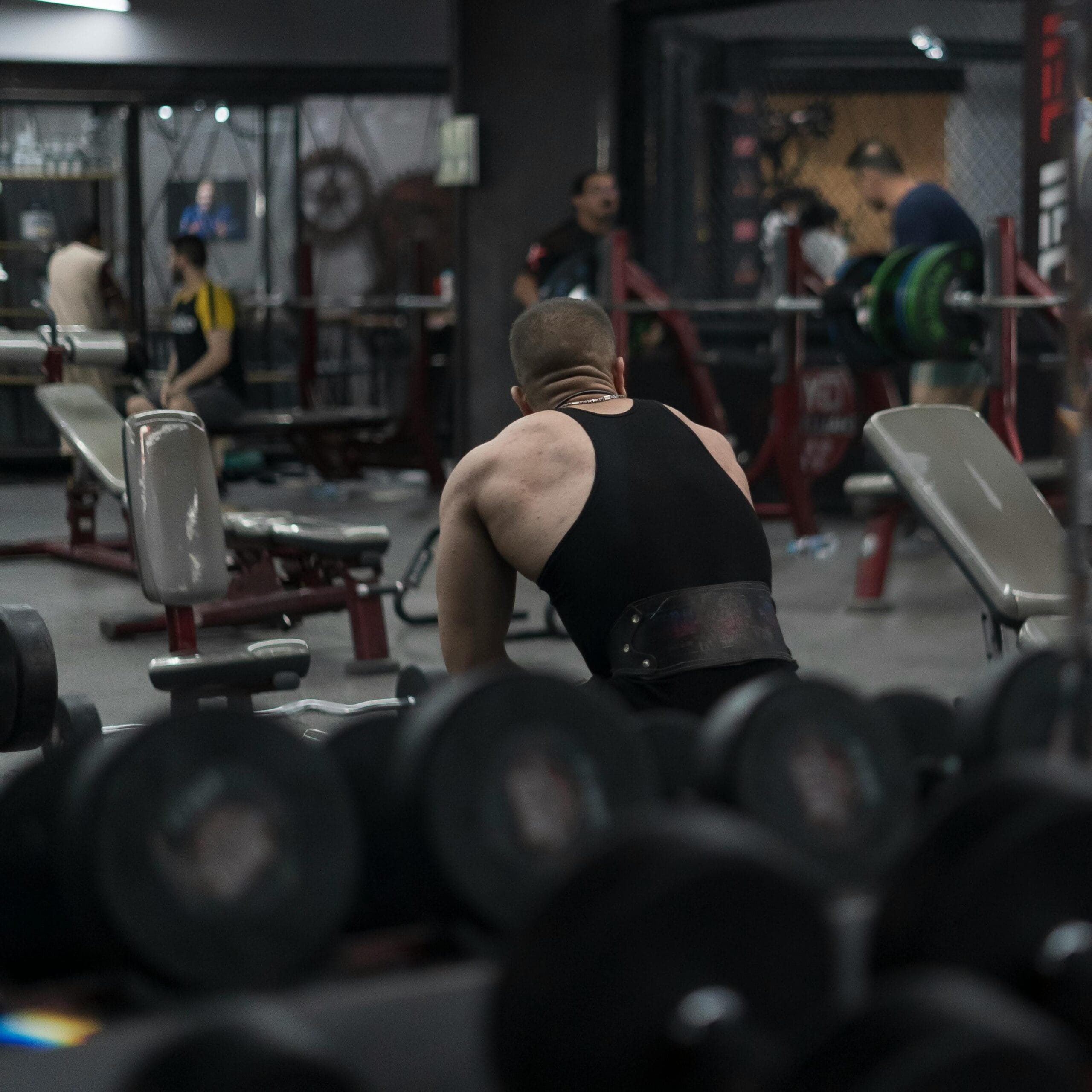 A muscular man with a black tank top working out in a gym with dumbbells visible.